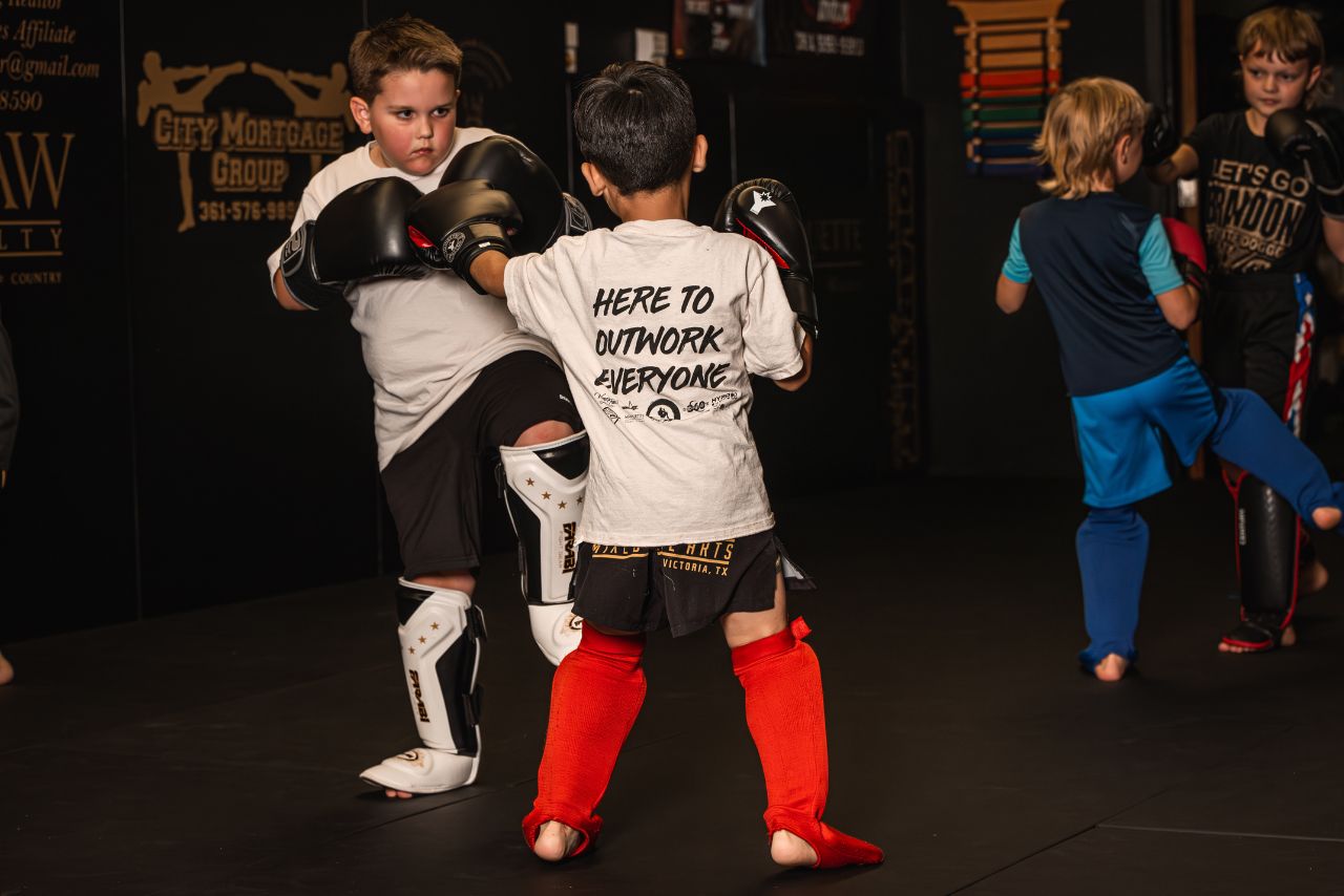 Two young boys practicing martial arts, wearing protective gear including gloves and shin guards