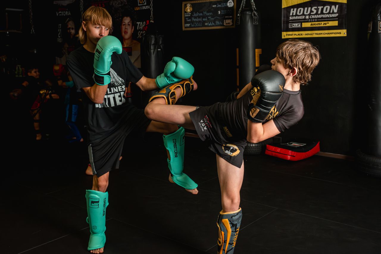 Two boys practicing kickboxing in a gym, wearing protective gear including gloves and shin guards.